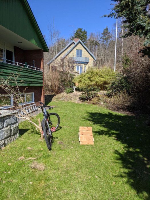 Mountainbike står på en gräsmatta bredvid en hembyggd träramphopp, med ett hus och trädgård i bakgrunden.