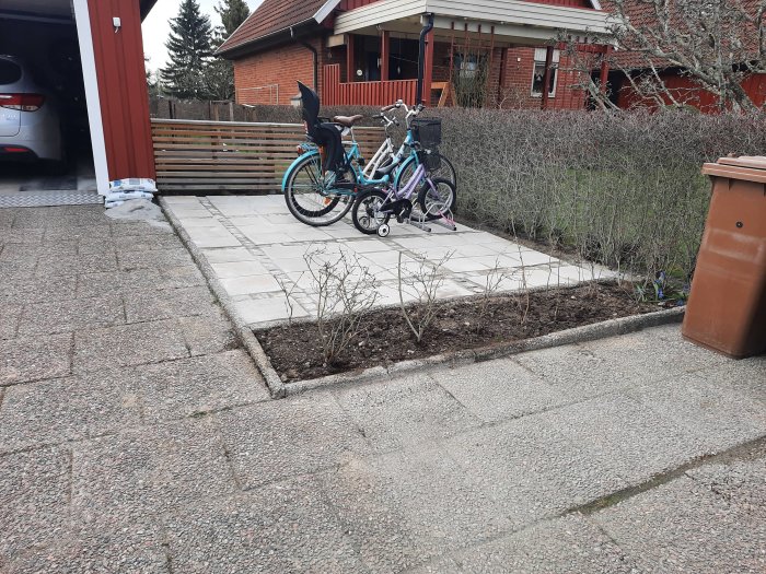 Uppfart i stenläggning med en vit plattlagd yta och en nyplanterad rabatt framför en röd tegelvilla med cyklar parkerade.