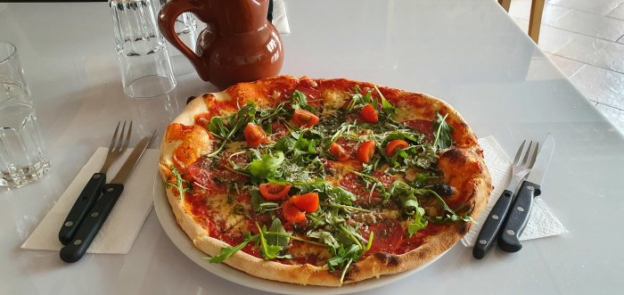 En vedugnsbakad pizza toppad med mozzarella, färsk ruccola och körsbärstomater, serverad på ett vitt bord med bestick.