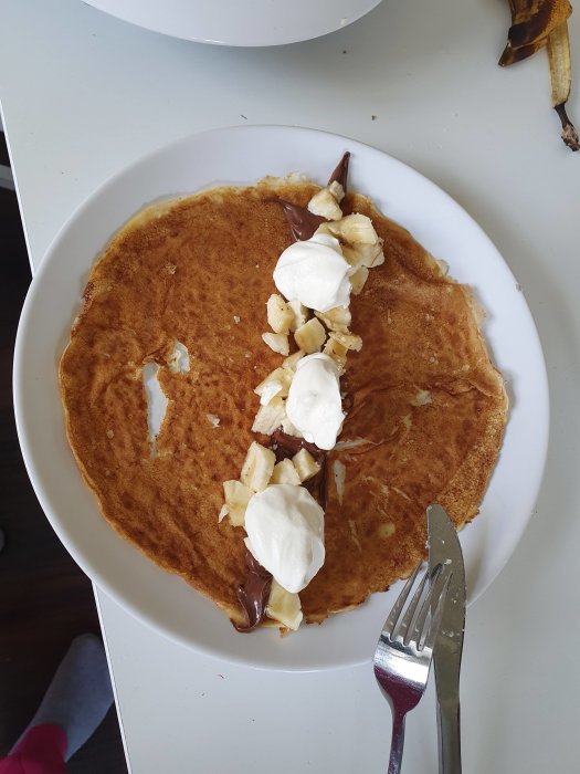 Pannkaka med Nutella, bananskivor och vispad grädde på en vit tallrik, med en gaffel vid sidan.