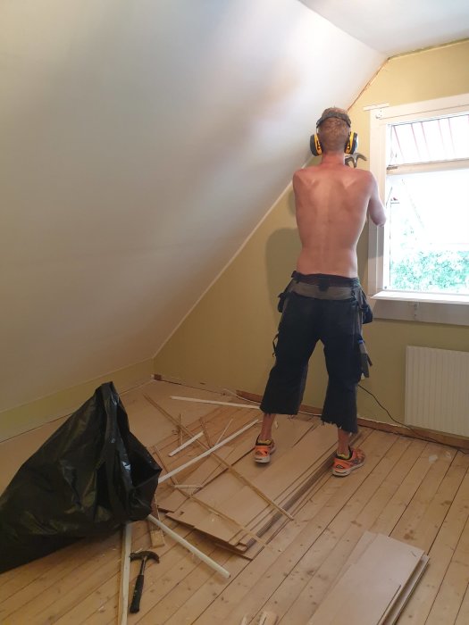 Person i hörselkåpor och verktygsbälte renovating ett rum med borttagna lister och utspridda byggmaterial.