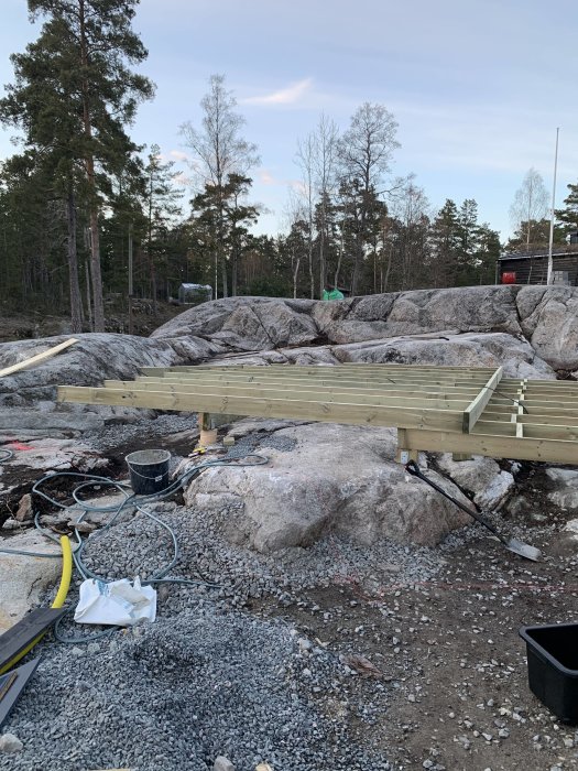 Byggplats med pågående arbete av trädäcksgrundläggning på klippterräng med skog i bakgrunden.