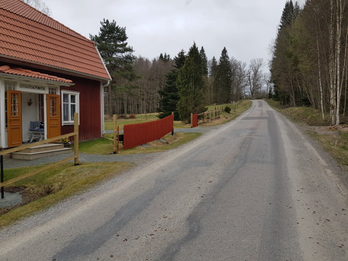 Röd trästaket vid landsväg nära rött hus med grön gräsmatta och skog i bakgrunden.