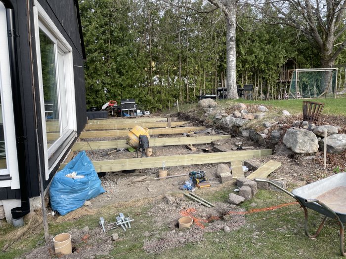 Byggprojekt med halvfärdiga plintar, byggmaterial och verktyg framför ett hus och trädgård.