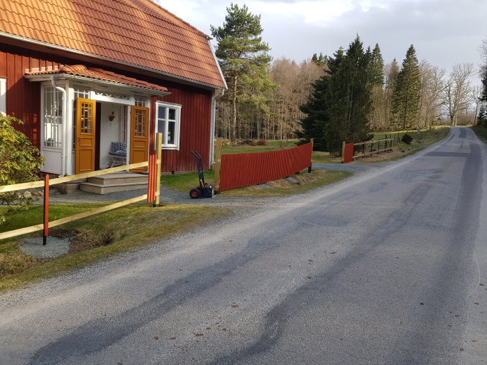 Rödmålat trästaket med nysatta spjälor utanför rött hus med gult dörrparti och handgräsklippare i förgrunden.