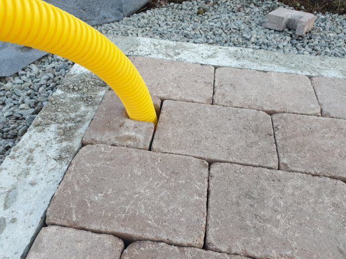 Nyinstallerade markplattor med ett anpassat uttag för ett gult dräneringsrör, intill grågrus och betongkant.