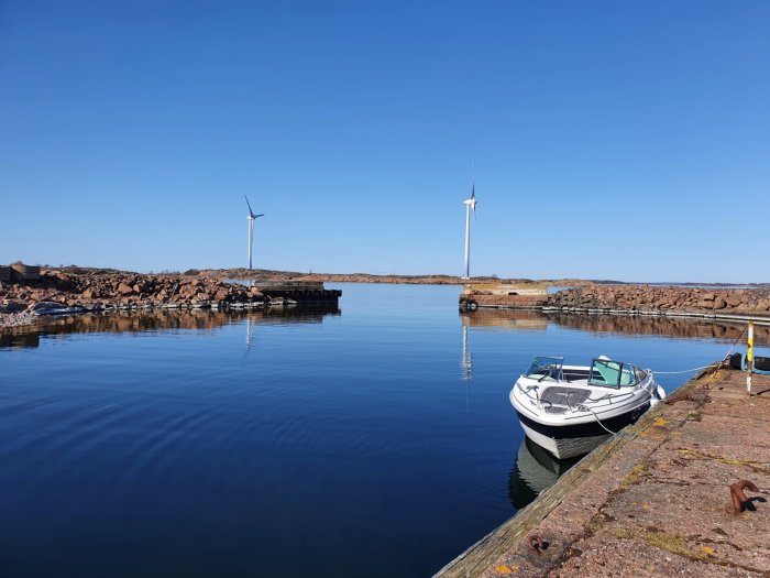 Motorbåt förtöjd vid kaj med vindkraftverk och klarblå himmel i bakgrunden.