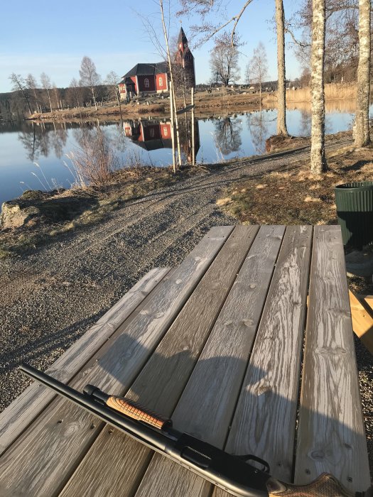 Gevär på träbrygga med spegling av röd stuga i vattnet i bakgrunden.