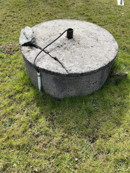 Bild på en betongbrunn med lock och en löst liggande kabel, i en blöt gräsmatta.