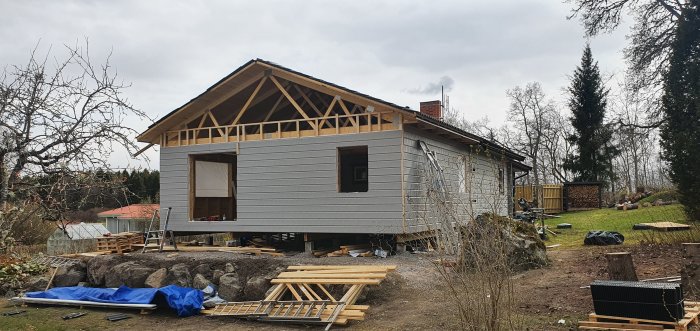 Halvfärdigt hus med trästomme och grå ytterpanel, byggmaterial spridda omkring.