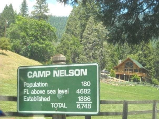 Skylt med texten "CAMP NELSON" visar befolkning och höjd över havet framför naturlandskap och en trästuga.