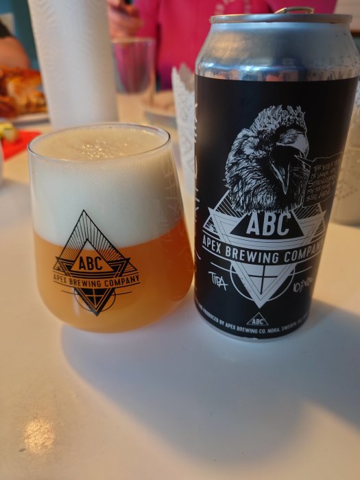 Ölglas med skum och ölburk från Apex Brewing Company på ett bord.