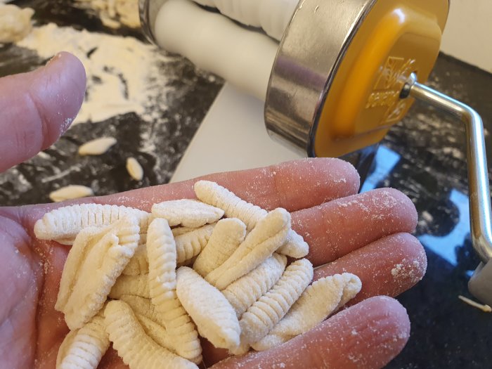 Hemmagjord cavatelli pasta på handflatan framför pastamaskin och mjölad yta.