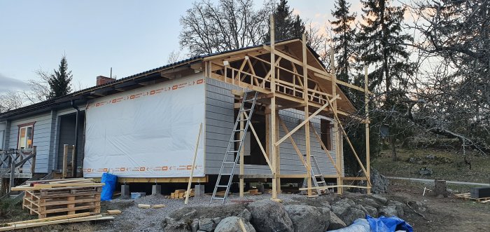 Hustillbyggnad under arbete vid skymning med träställning, säkerhetsstängsel och byggmaterial.