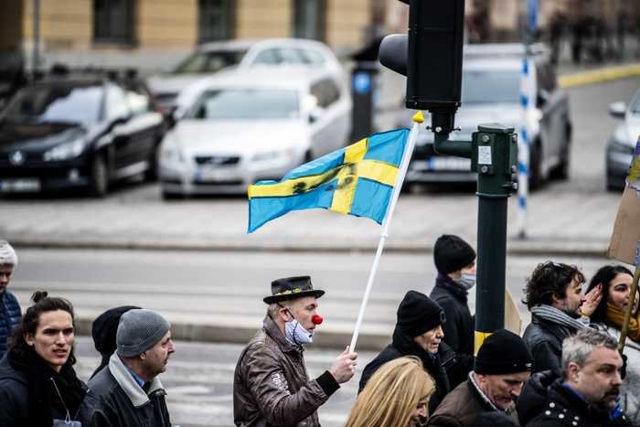 Person i clownsmink och hatt viftar med svensk flagga bland andra människor.