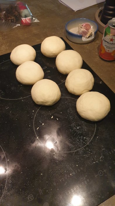 Runda degbollar på mörk bakplåt redo för jäsning, flugit mjöl, vitlöksskål och läskburk i bakgrunden.