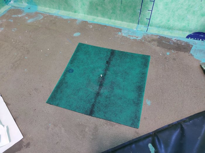 Grön självhäftande våd ligger centralt på ett betonggolv redo för installation över avloppet i ett pågående byggprojekt.