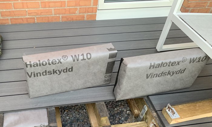 Två förpackningar av Halotex vindskydd på en veranda med grå trätrall och tegelvägg.