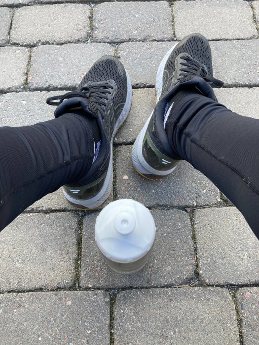 Person i löparskor och tights sittandes på stenläggning med en vattenflaska framför sig.