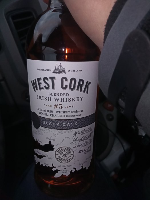 Flaska West Cork Blended Irish Whiskey hållen i någons knä i ett fordon.