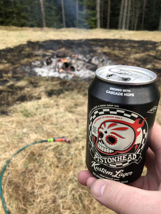 Hand håller ölburk av märket "Pistonhead Kustom Lager" framför en eld och en vattenslang i naturen.