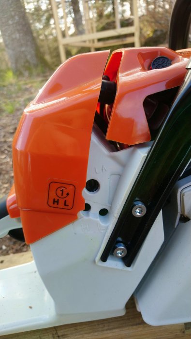 Detaljbild på en motorsåg med skruvhål och märkningen 'HL' på en orange och vit kropp.