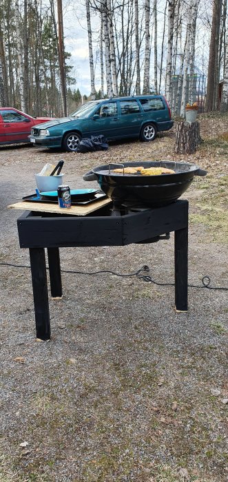 Hemmagjord grillbåt av spillvirke med kött på grillen, plywoodskiva och ölburk framför bilar och björkar.