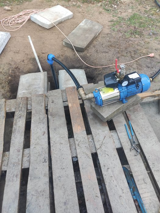 Vattenpump och kopplingar monterade ovanför en brunn, med slangen nedförd i röret och verktyg på marken.
