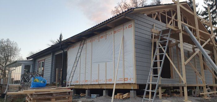 Byggprojekt av ett hus vid skymning med ny spikläkt och skyddsplast på ena sidan, trästolpar och byggnadsställning syns.