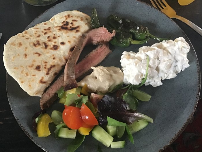 Valborgsmiddag med flankstek, potatissallad, hummus, stekpannebröd, stekta padrones och sallad på en tallrik.