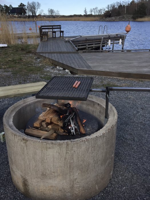 Nyinvigd grillplats vid sjö med brygga, eld i betongring och korv på grillgallret.