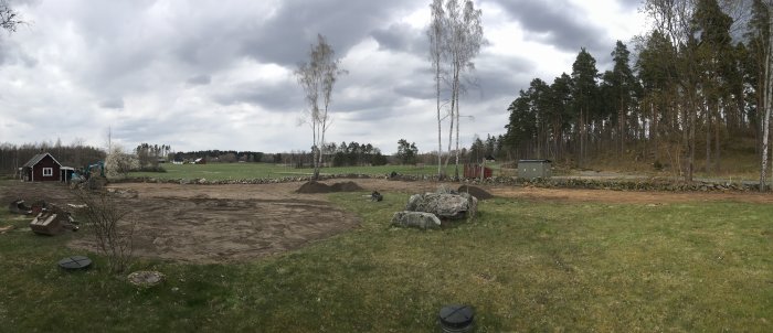 Panoramavy av en trädgård under återställning, med blottad jord, stenar och maskindelar.