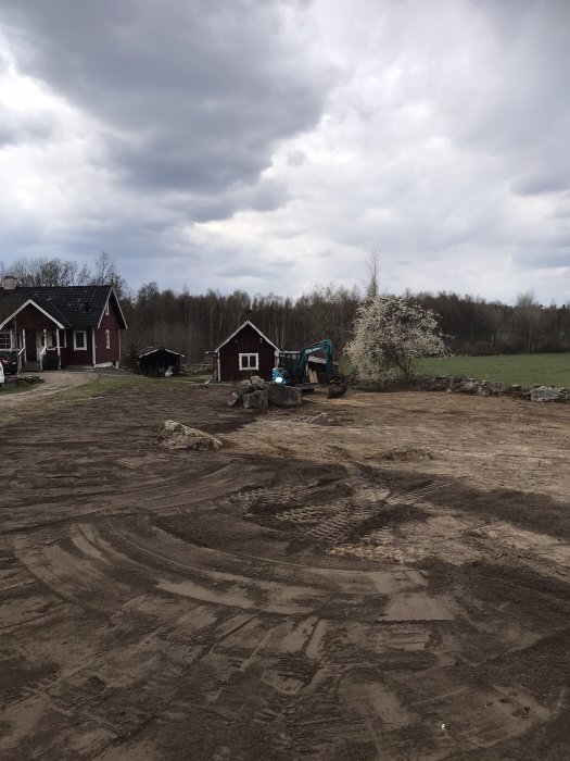 En grävare på en jordyta med spår och röjning framför ett rött hus och blommande träd.