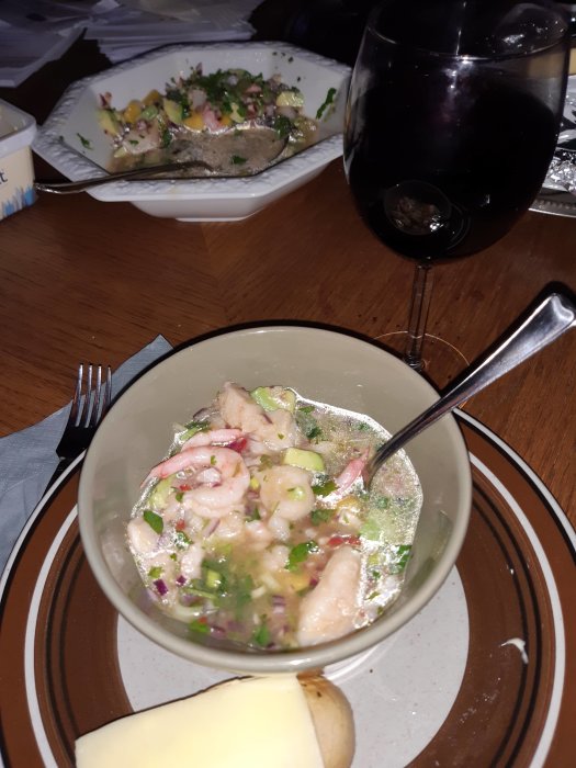 En skål med ceviche framför en smörgåstårta och ett glas rödvin på ett matbord.