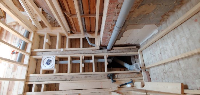 Pågående badrumsrenovering med nyinstallerade avloppsrör och hålband synliga mellan träreglarna i golv och vägg.