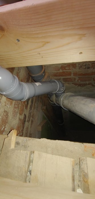 Nyinstallerade avloppsrör i grått under en träbjälke, med band för fastsättning synlig.