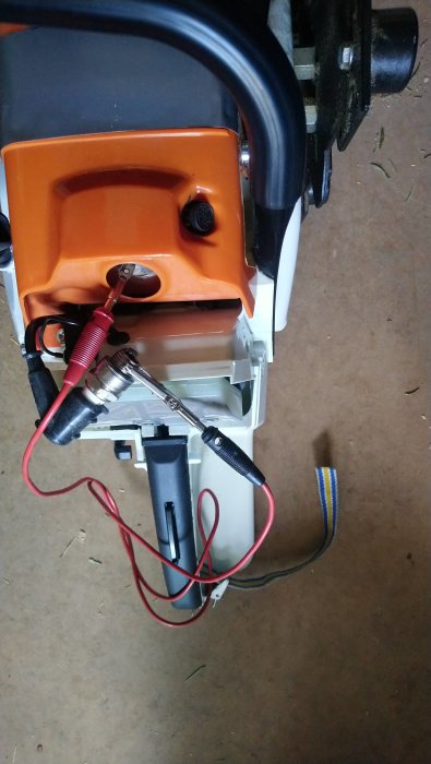 Tändstift testat mot motorblock med röd och svart kabel visande gnistbildning, bredvid delvis monterad maskindel.