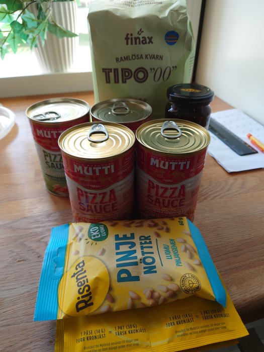 Konservburkar med tomatsås, påse med mjöl och paket av pinjenötter på ett köksbord.