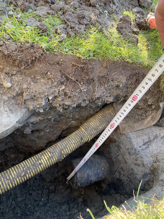 Måttband visar djupet på 50cm i en grävd grop där ett gult anslutningsrör partiellt täcks av jord.