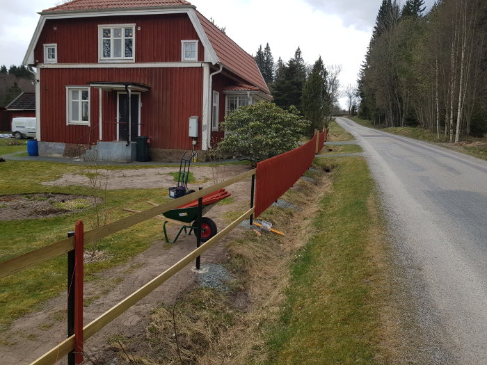 Nyuppsatt rött staket längs en slänt vid infart, med skottkärra och redskap i förgrunden och rött hus i bakgrunden.