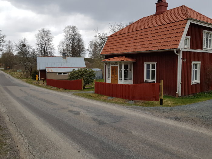 Rött trähus med nytt staket och stödmur vid infart, flackare slänt nära vägen.