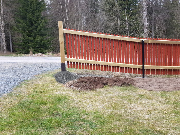 Nybyggt rött staket längs en grässlänt och en liten ny stödmur av sten vid sidan av en grusväg.