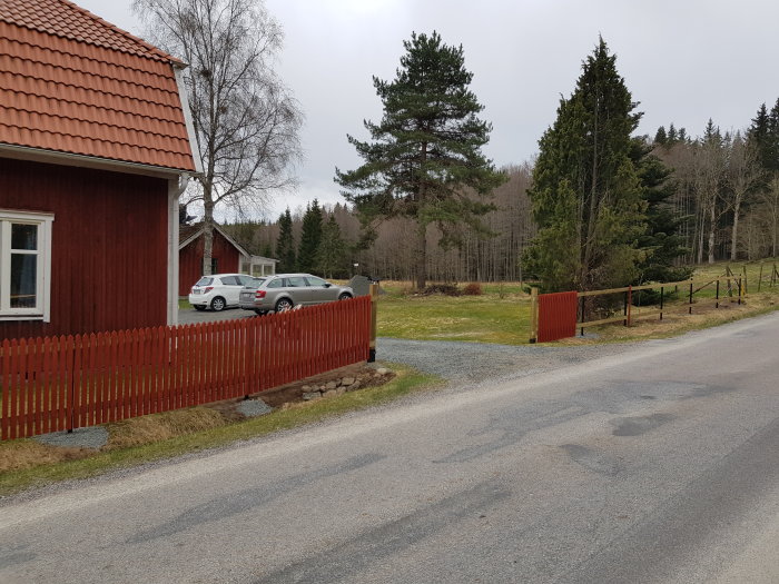 Nybyggd stödmur längs grusväg med nyligen uppställt rött staket och två parkerade bilar i bakgrunden.