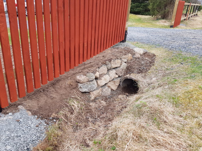 Nybyggd stödmur av sten intill slänt och staket vid infart, jord och grus synliga.