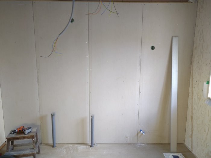 Badrumsvägg med nyligen monterade gipsskivor, synliga rör och elledningar samt ett verktyg på en pall.