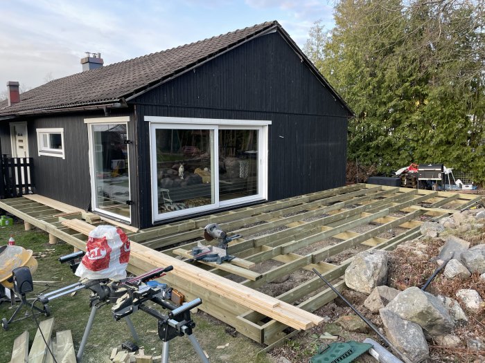 Nybyggd trägrund till terrass framför svart trähus med verktyg och material synliga.