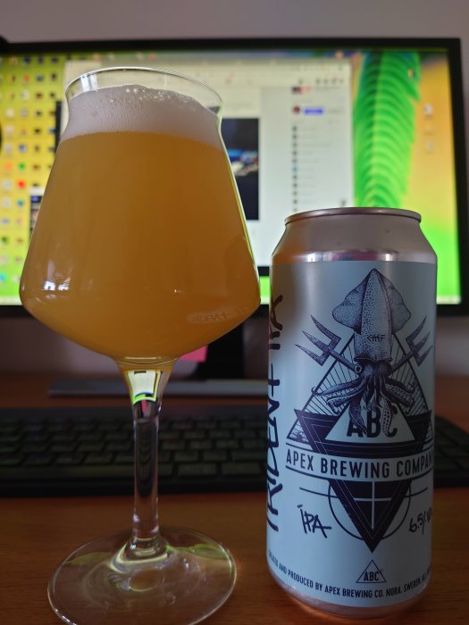 En ölglas fyllt med hazy IPA står framför datorskärm med öppet diskussionsforum, bredvid en ölburk från Apex Brewing.