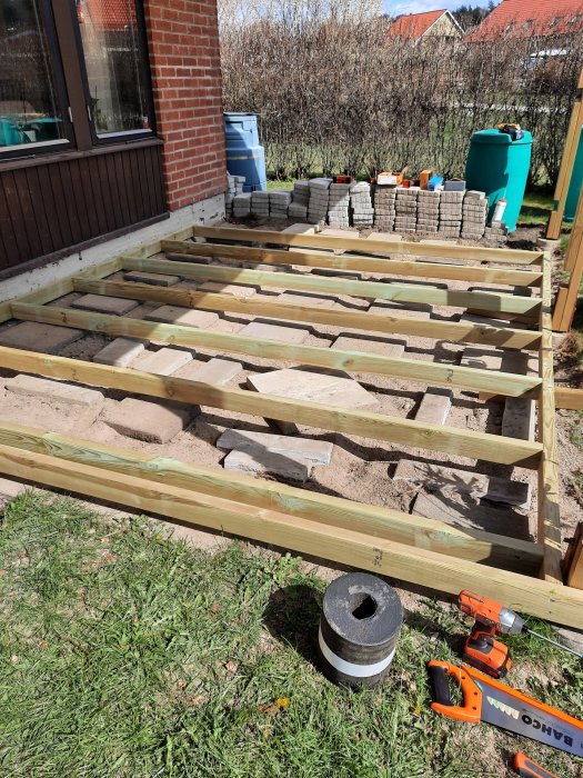 Påbörjad konstruktion av altanens regelverk med träbalkar monterade på stenplattor, verktyg synliga.