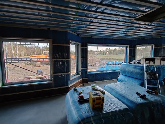Interiör av ett hus under konstruktion med synliga isoleringsmaterial och stora oskyddade fönster.