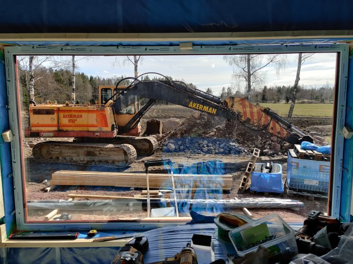 Utsikt genom ett stort fönster med en grävmaskin som flyttar jord och byggmaterial framför.